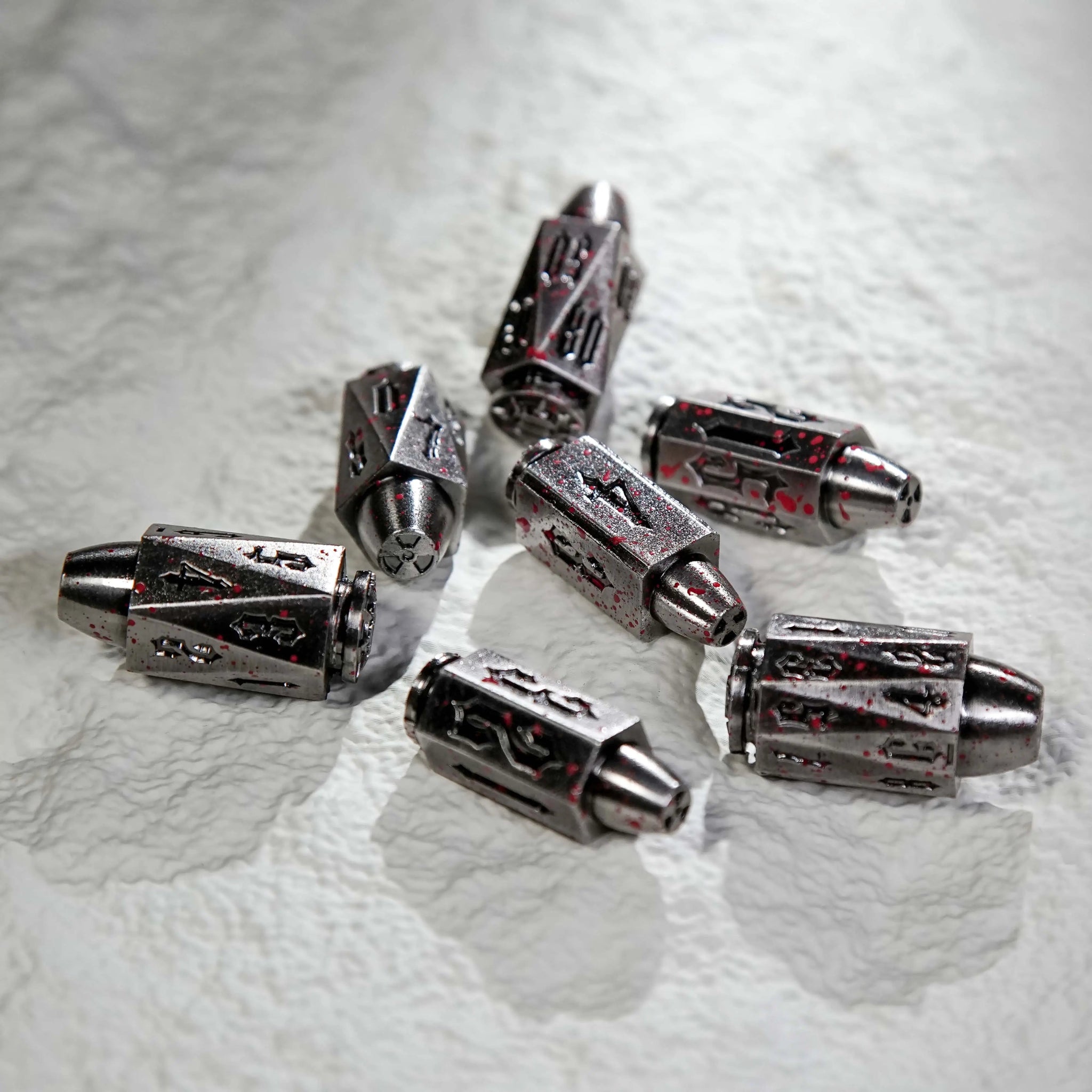 A cool blood-stained polyhedron DND metal bullet dice set is scattered on a white table. Bullet-shaped RPG dice | Bear Dice
