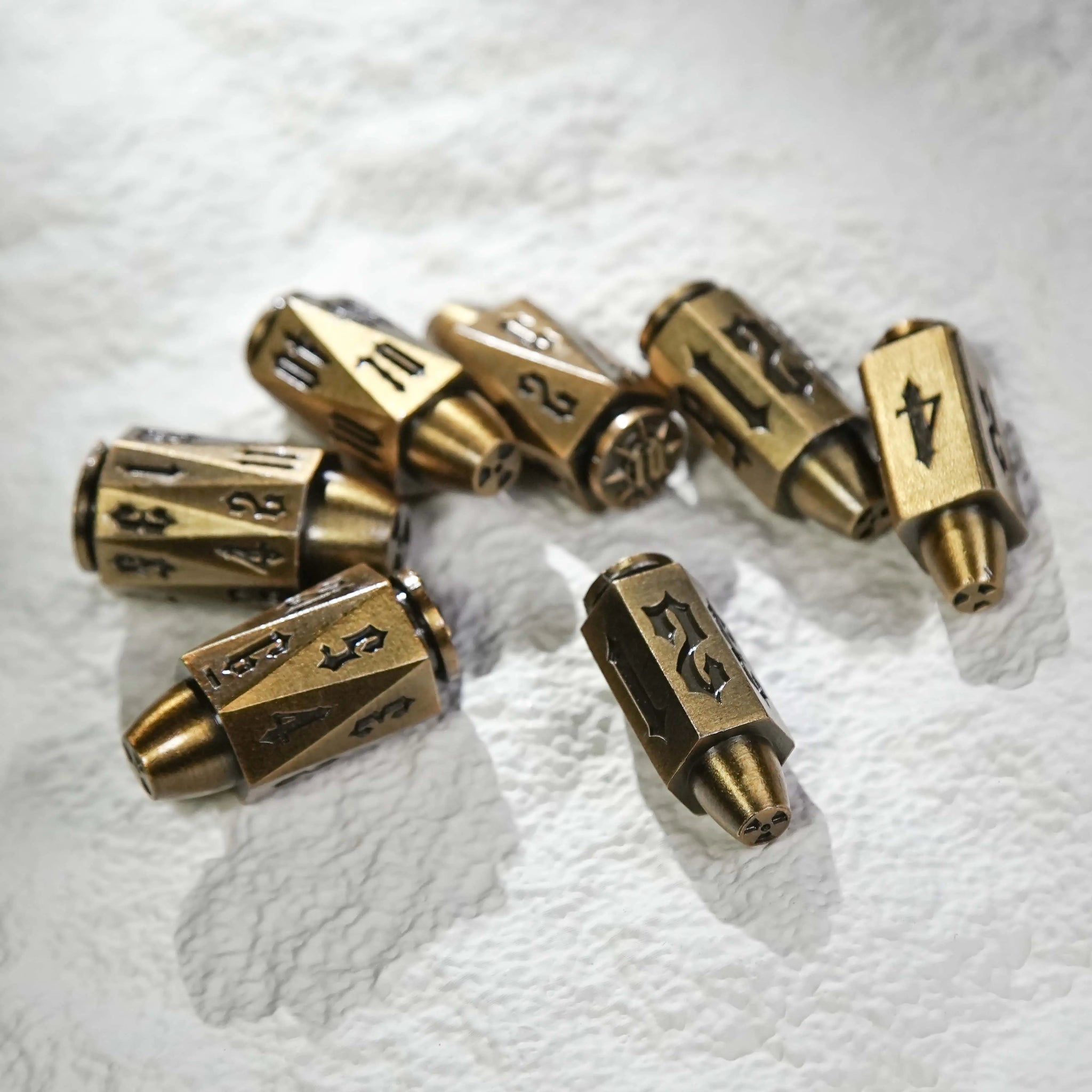 Cool antique gold polyhedron DND metal bullet dice set scattered on a white table, bullet-shaped RPG dice | Bear Dice