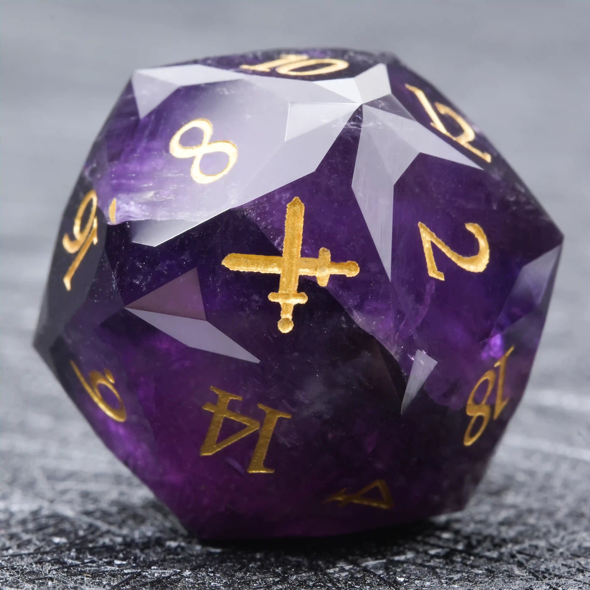 The facet of this multi-faceted natural amethyst D20 die is engraved with a double swords pattern, and the numbers are gold. | Bear Dice